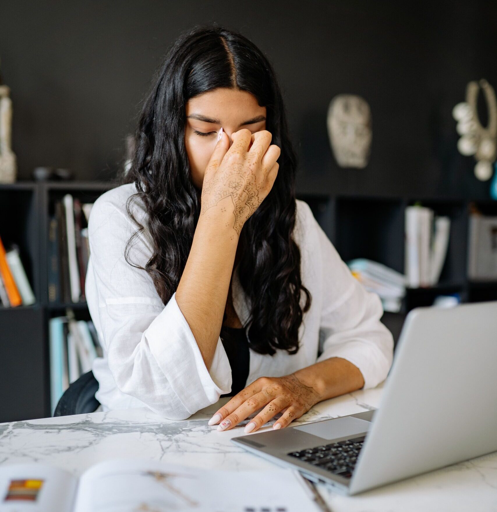 A woman feeling stressed while working on a laptop in an office setting with documents.